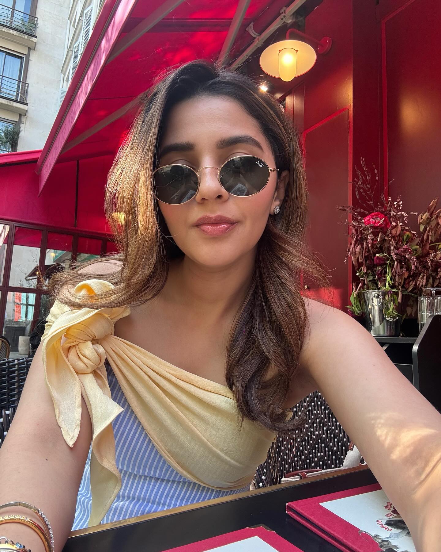 A woman wearing an off-shoulder yellow and white striped top with a side tie, paired with sunglasses and sitting at a table with red umbrellas in the background.