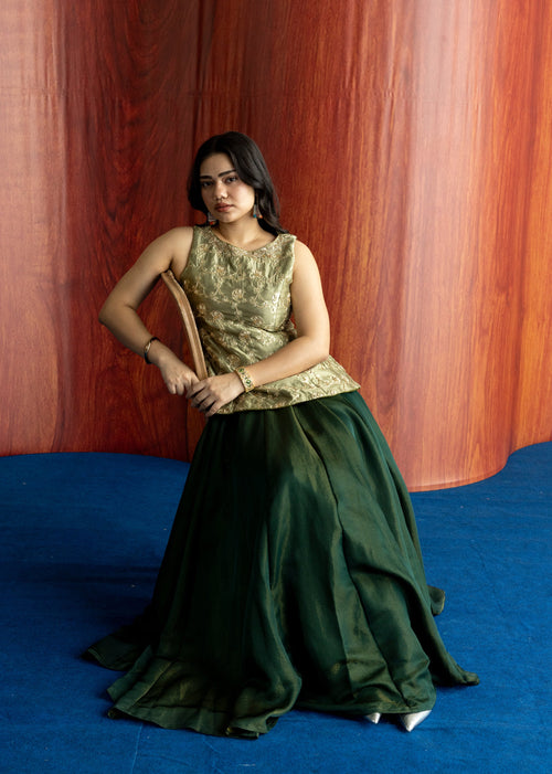 A woman wearing a sleeveless formal dress with a gold patterned bodice and a dark green skirt, standing on a blue floor against a curved wooden backdrop.