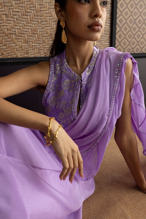 A woman wearing a lavender purple sleeveless kurta with gold floral embroidery and a matching sheer draped fabric, styled with gold jewelry in an indoor setting.